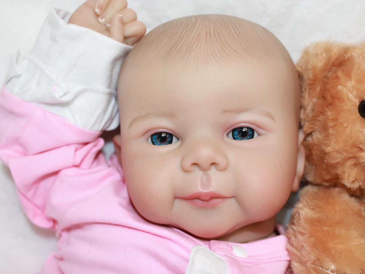 A baby doll rests on a white blanket beside a soft teddy bear, creating a cozy and playful scene.