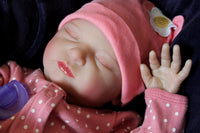 Thumbnail for A baby doll resting on a soft purple blanket, showcasing its delicate features and bright clothing.