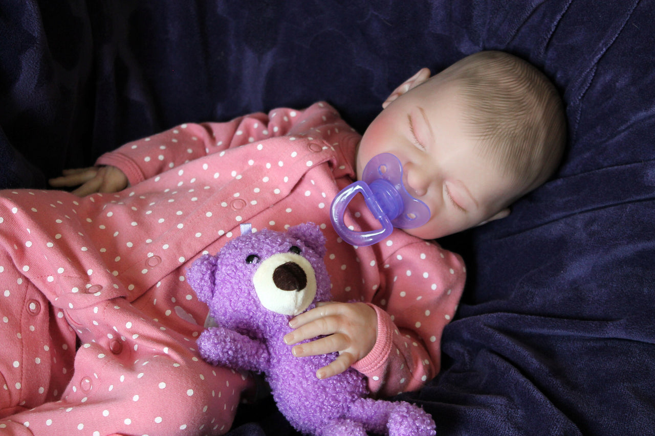A baby doll lying on a cozy purple blanket, emphasizing its sweet face and playful attire.