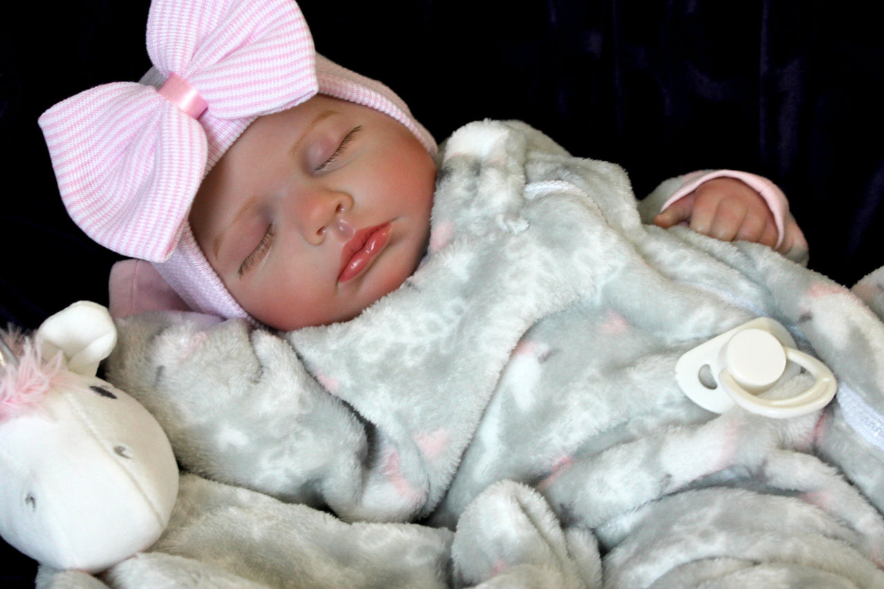 A newborn baby girl dressed in gray with a pink bow, sleeping adorably on a cozy surface.