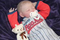 Thumbnail for A baby dressed in a baseball uniform rests on a vibrant purple blanket, exuding cuteness and charm.