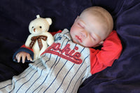 Thumbnail for A baby dressed in a baseball uniform rests on a vibrant purple blanket, exuding cuteness and charm.
