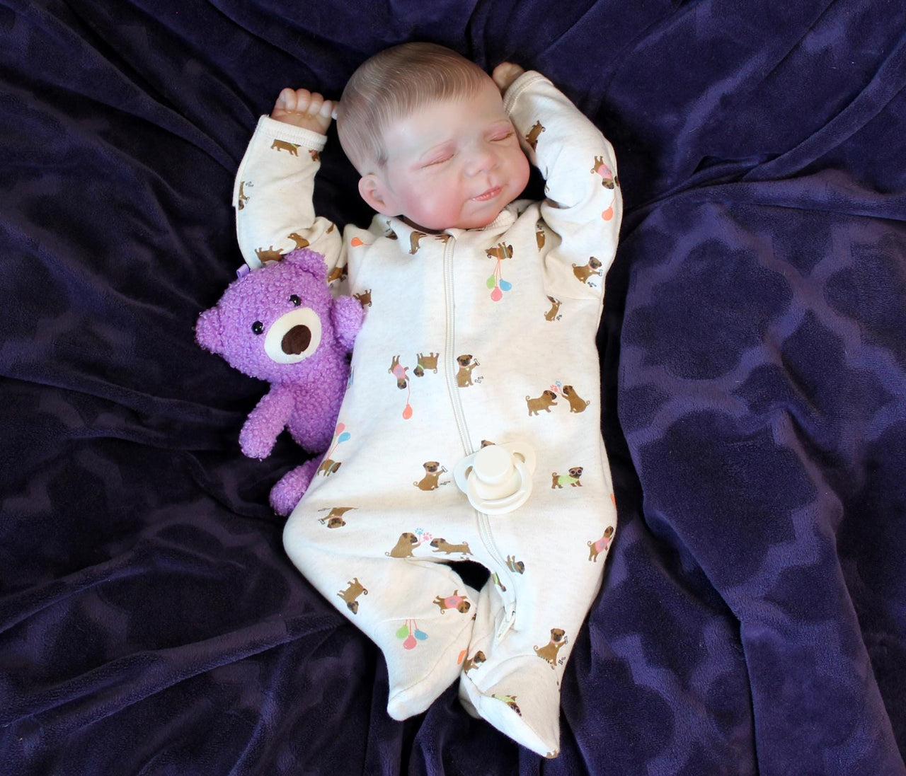 A baby in a onesie lies on a soft purple blanket, looking peaceful and content.