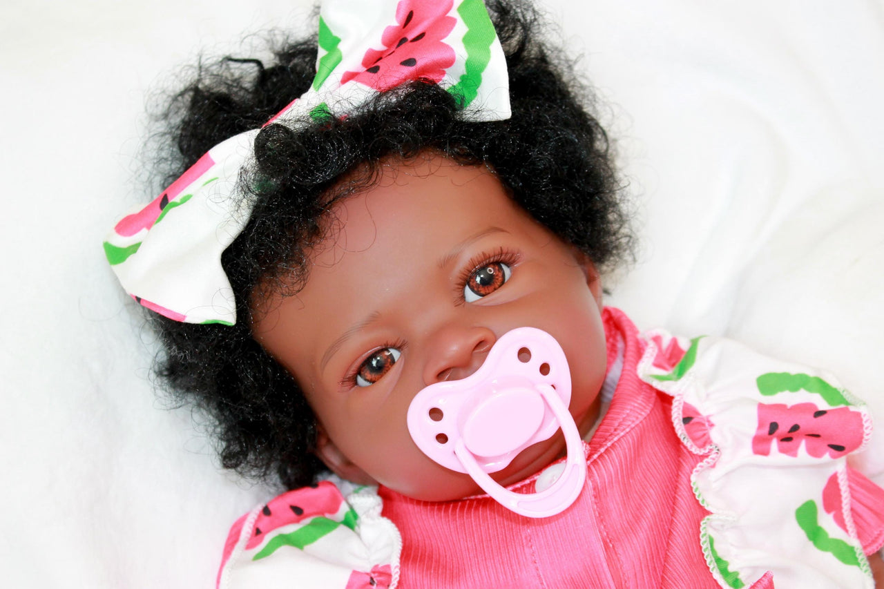 A black baby doll positioned on a white bed, highlighting its charming design against a clean, bright background.