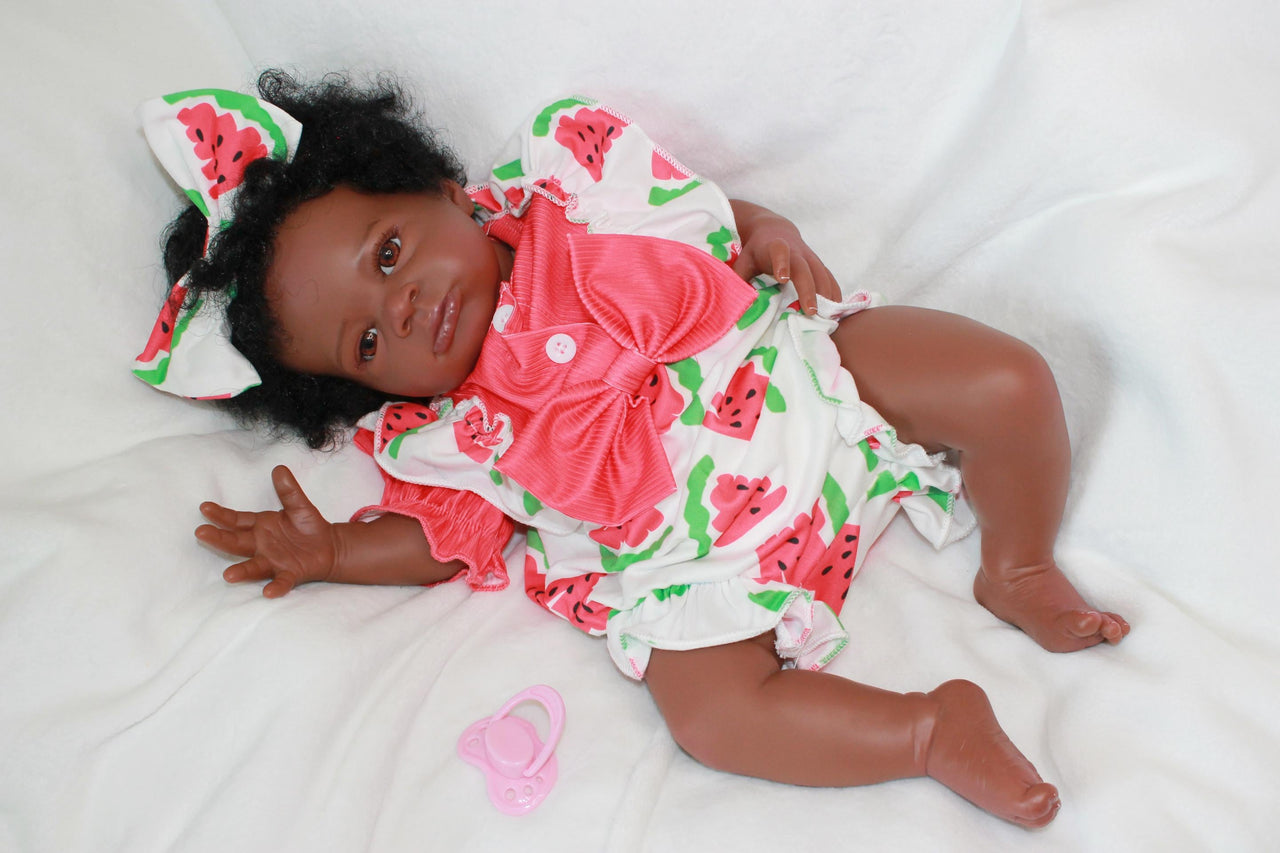A black baby doll positioned on a white bed, highlighting its charming design against a clean, bright background.