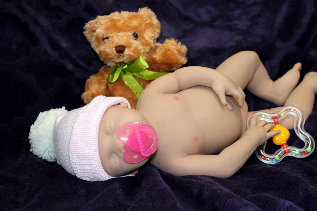 Full Silicone Doll baby doll lies on a vibrant purple blanket, accompanied by a charming teddy bear, evoking a sense of comfort and joy.