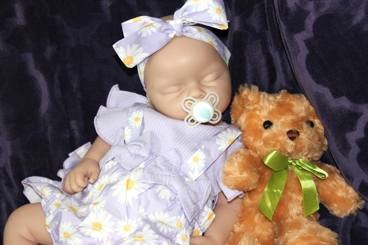 A baby doll wearing a purple dress, holding a teddy bear, sits on a soft surface, showcasing its charming features.