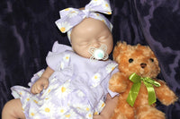 Thumbnail for A baby doll wearing a purple dress, holding a teddy bear, sits on a soft surface, showcasing its charming features.