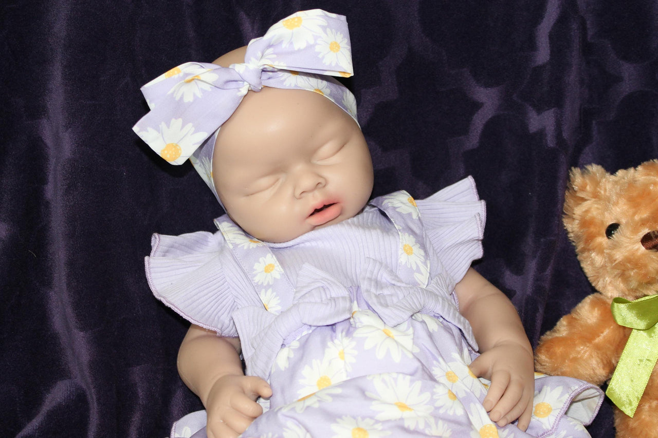 A baby doll wearing a purple dress, holding a teddy bear, sits on a soft surface, showcasing its charming features.
