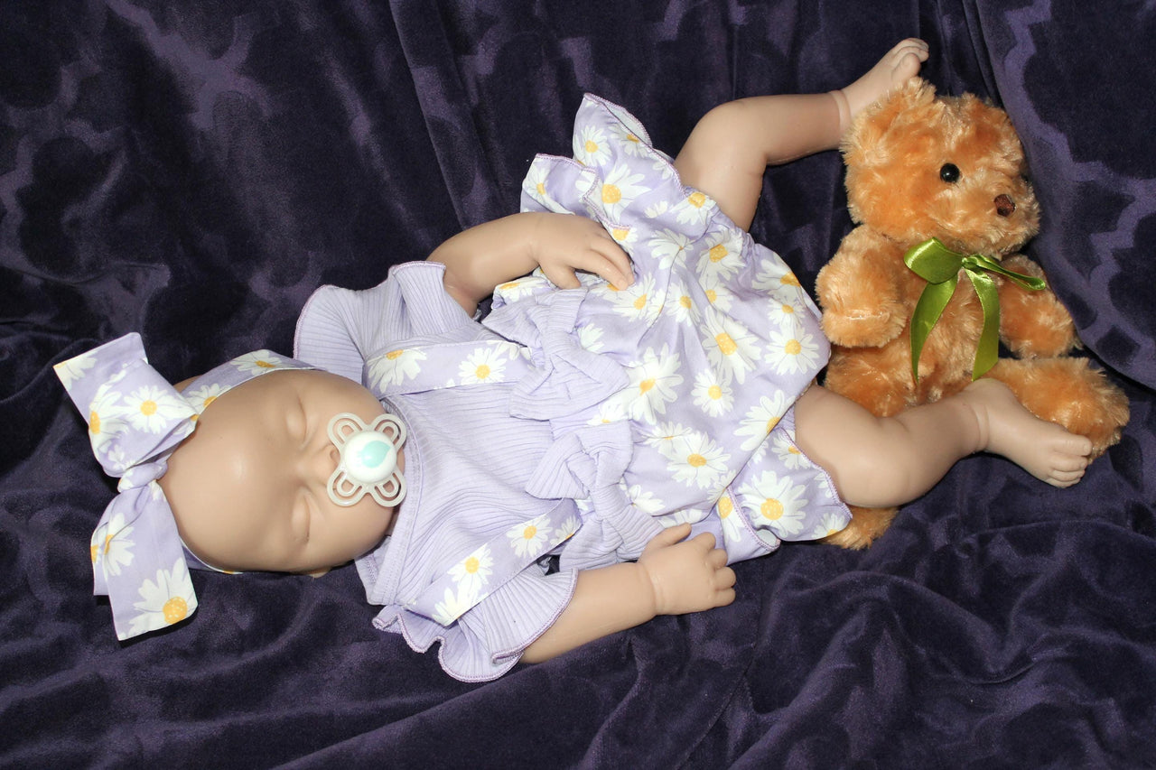 A baby doll wearing a purple dress, holding a teddy bear, sits on a soft surface, showcasing its charming features.