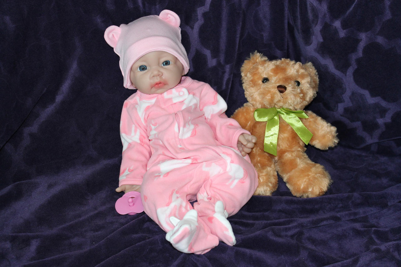 A baby doll holding a pacifier is placed next to a plush teddy bear on a neutral background.