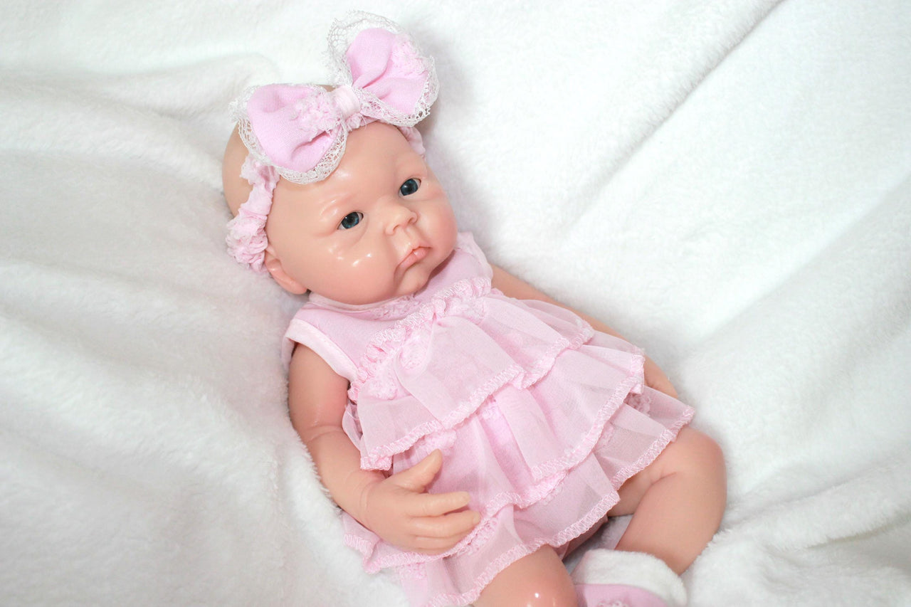 A baby doll in a pink dress and headband, showcasing a cheerful expression and delicate features.