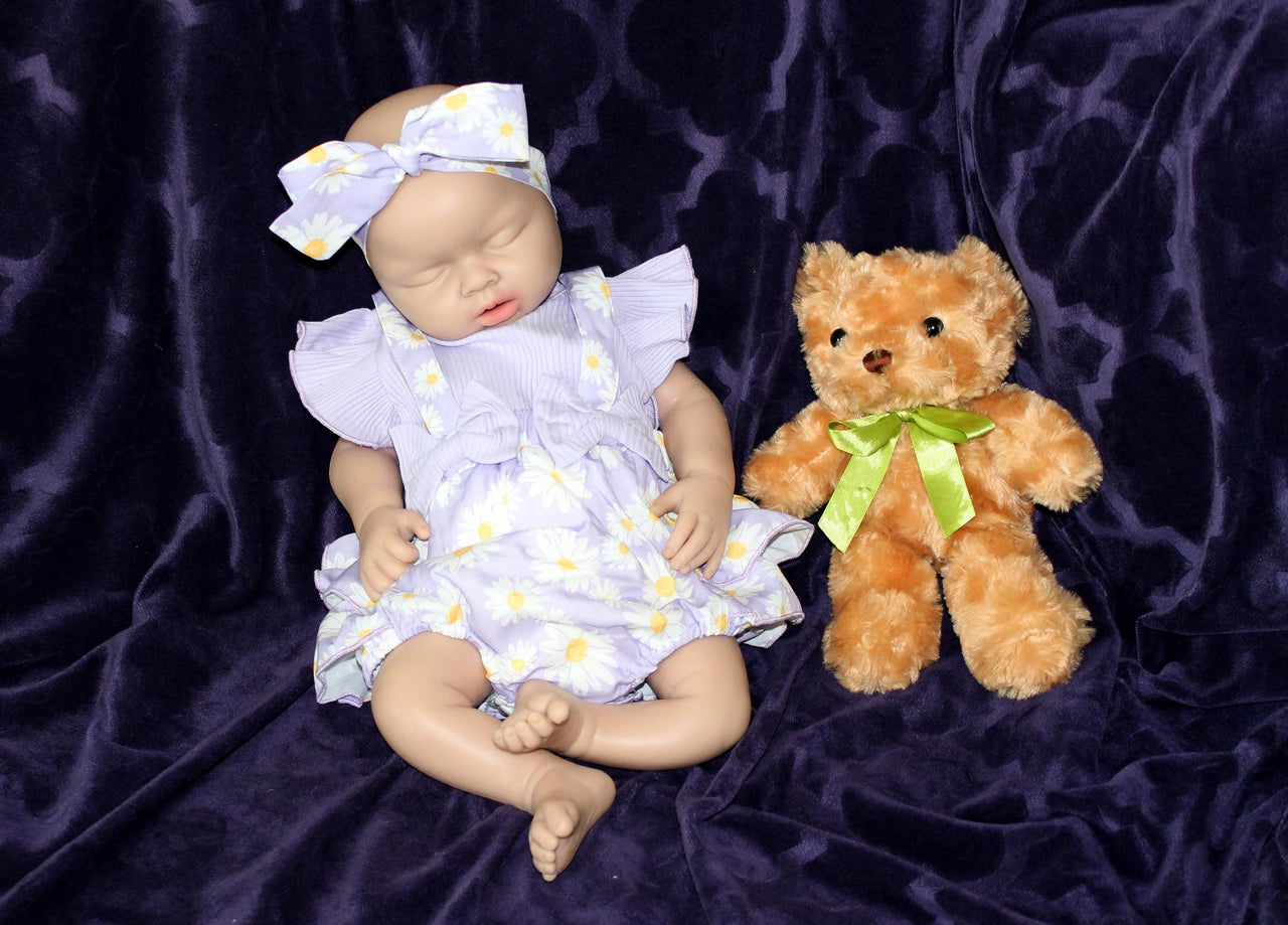 A baby doll wearing a purple dress, holding a teddy bear, sits on a soft surface, showcasing its charming features.