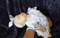 Thumbnail for A baby doll wearing a purple dress, holding a teddy bear, sits on a soft surface, showcasing its charming features.