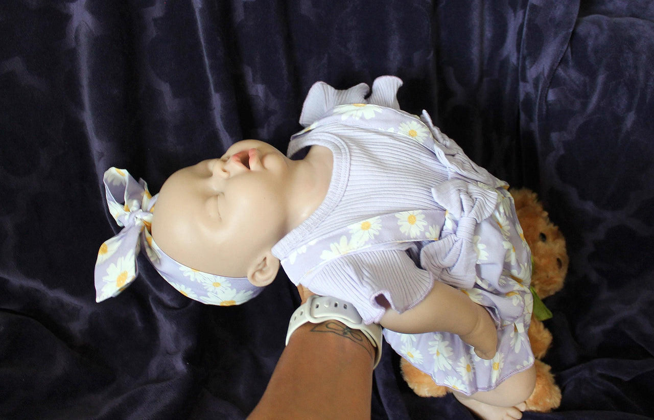A baby doll wearing a purple dress, holding a teddy bear, sits on a soft surface, showcasing its charming features.
