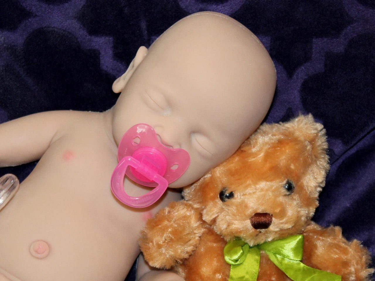 Full Silicone Doll baby doll rests on a soft purple blanket beside a cuddly teddy bear, creating a serene and playful scene.