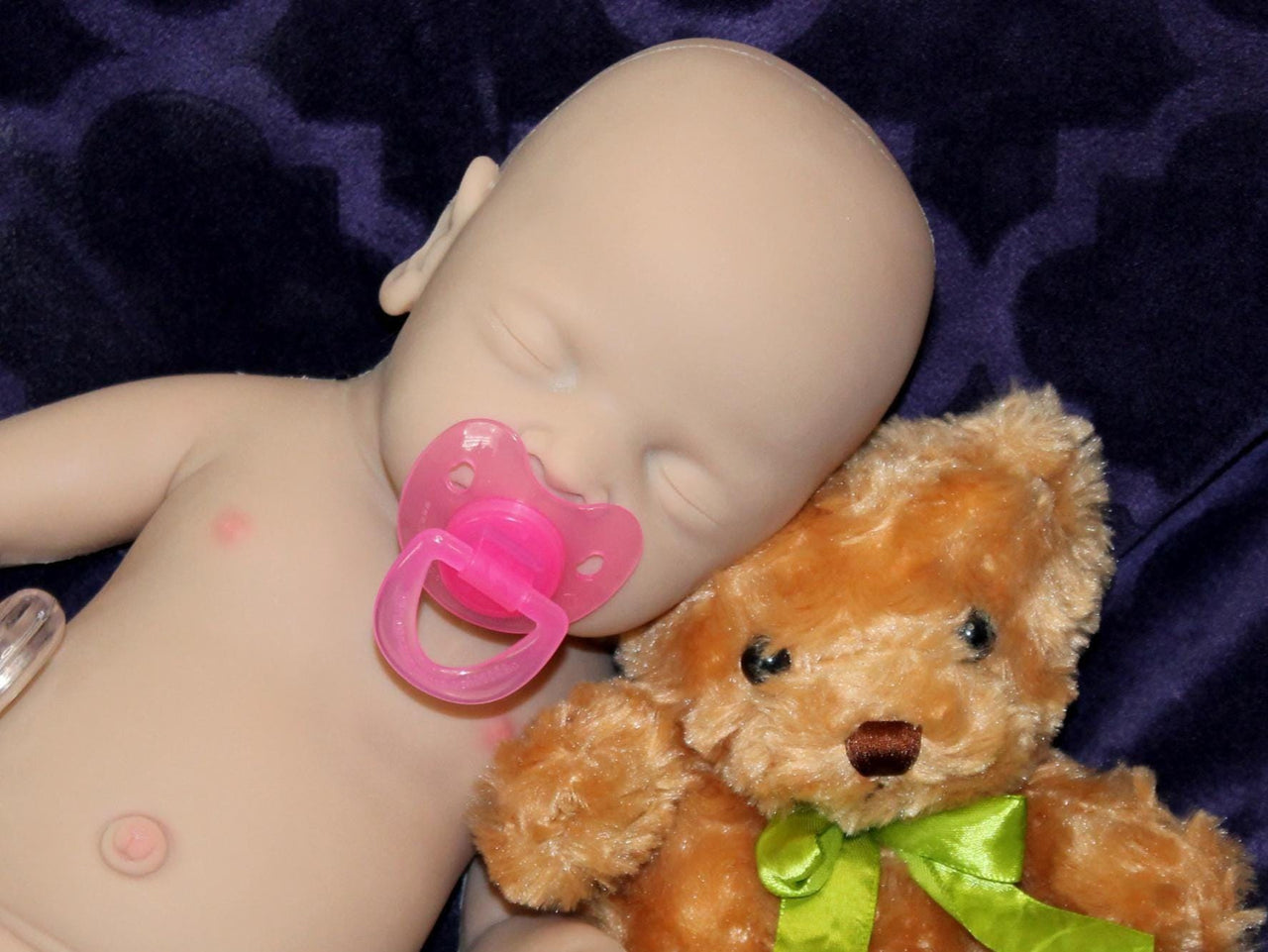 Full Silicone Doll baby doll rests on a soft purple blanket beside a cuddly teddy bear, creating a serene and playful scene.