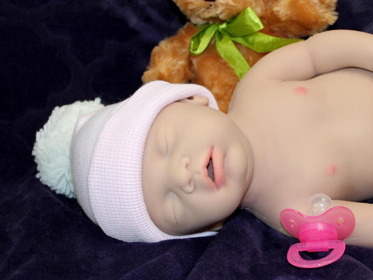 Full Silicone Doll baby doll is positioned on a purple blanket, next to a teddy bear, illustrating a delightful and cozy atmosphere.