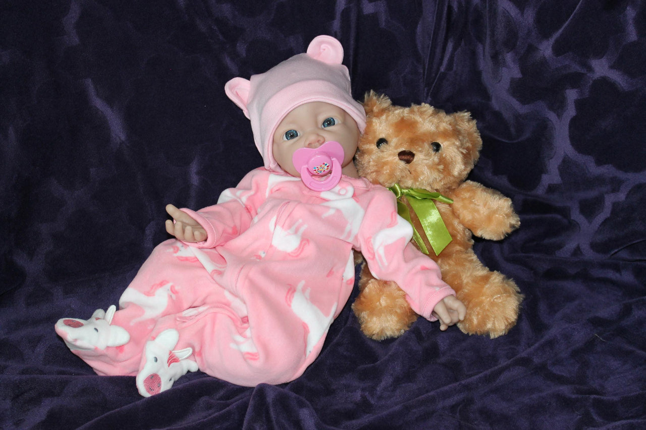 A baby doll with a pacifier sits beside a soft teddy bear on a light-colored surface.