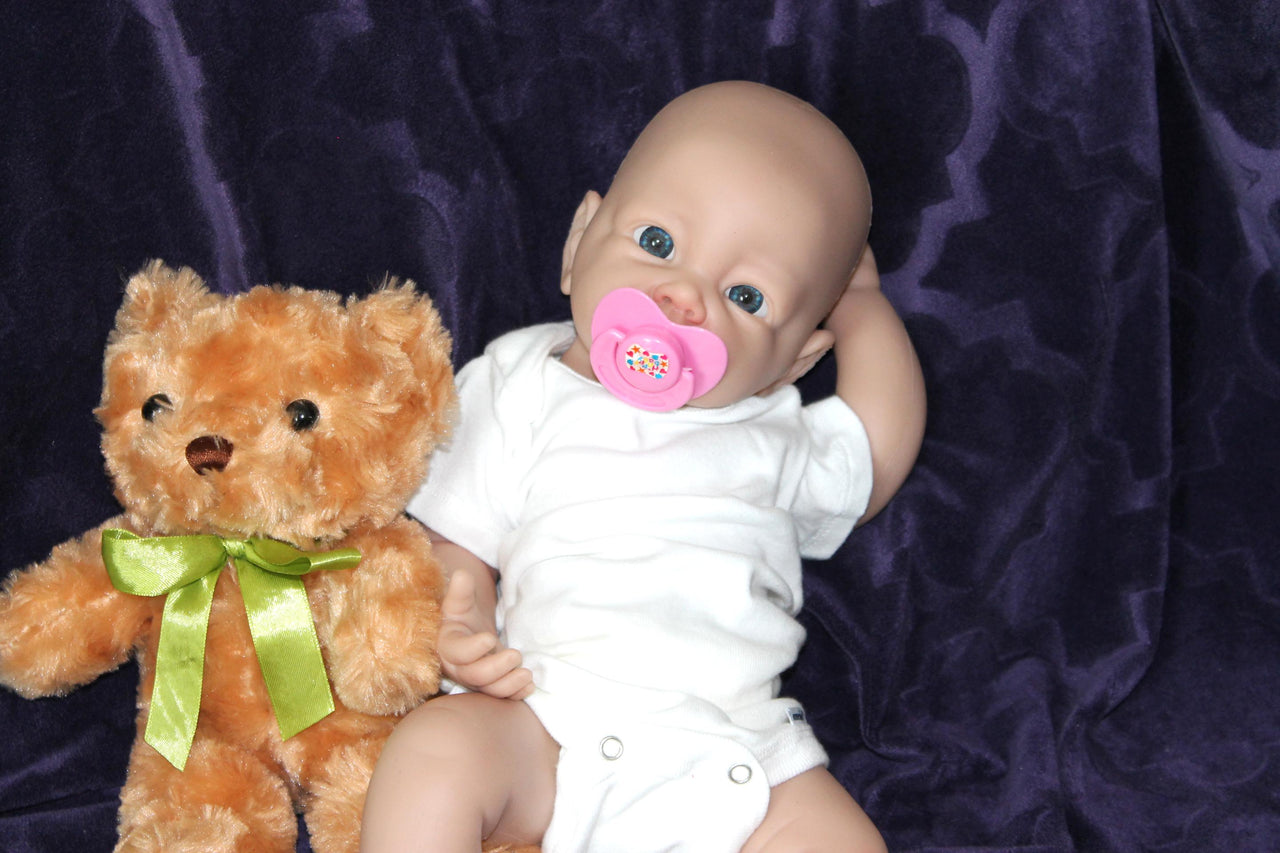 A baby doll holding a pacifier is placed next to a plush teddy bear on a neutral background.