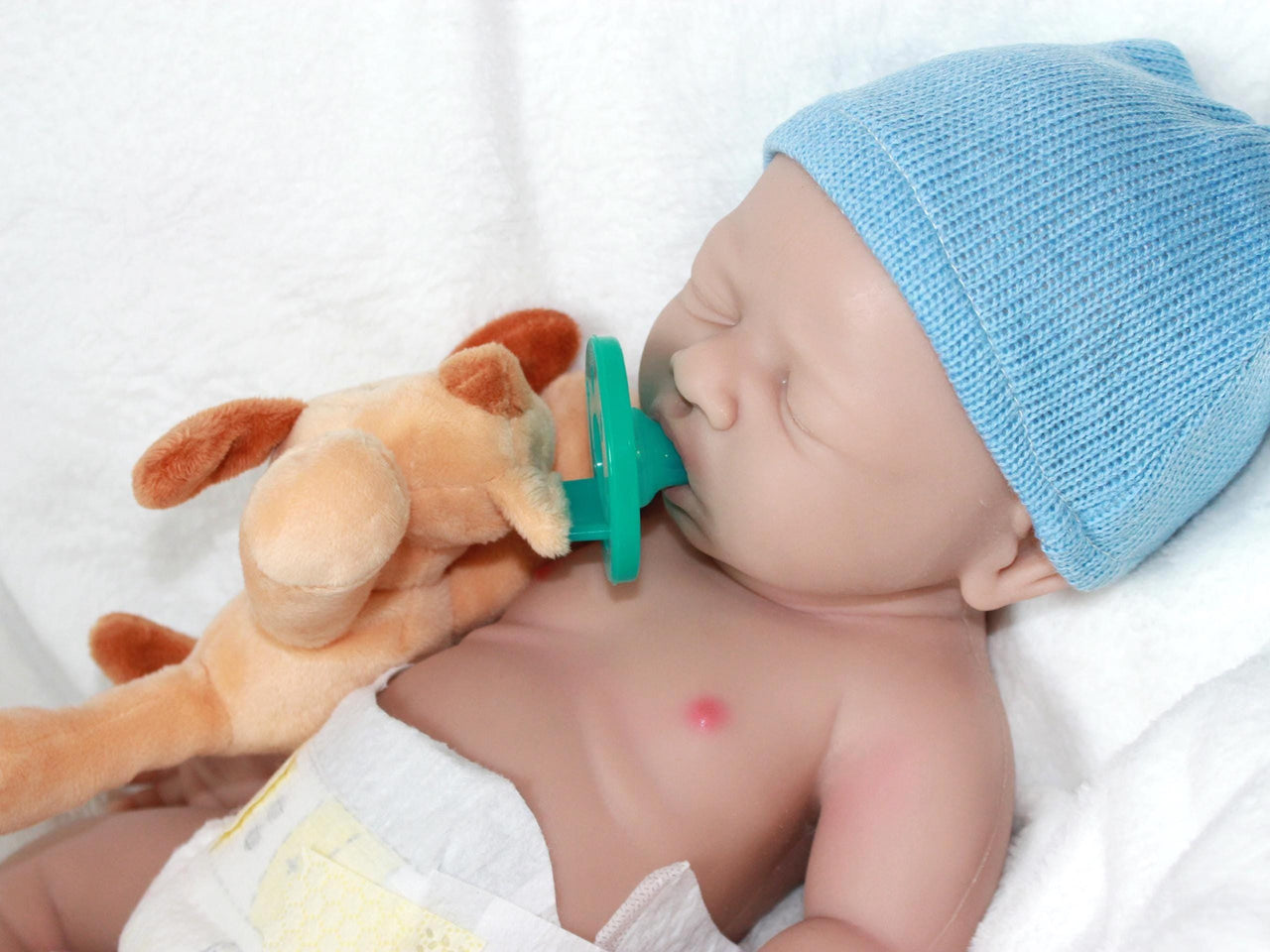 A baby doll wearing a blue hat is resting on a soft white blanket.