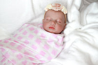 Thumbnail for A baby doll resting on a neatly made bed, surrounded by soft blankets and pillows.