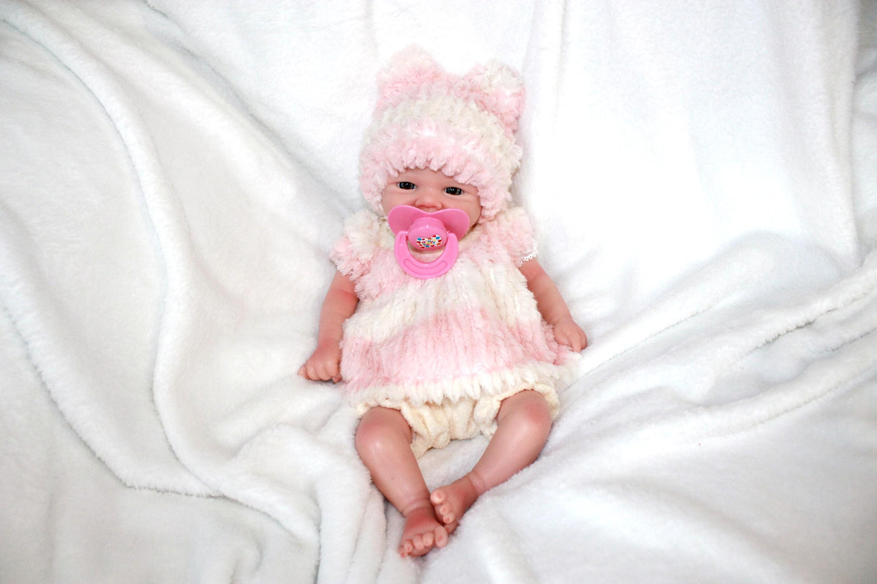 A baby doll dressed in a pink hat, featuring a pacifier, lying on a cozy blanket, highlighting its adorable design.