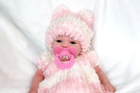 Thumbnail for A baby doll dressed in a pink hat, featuring a pacifier, lying on a cozy blanket, highlighting its adorable design.