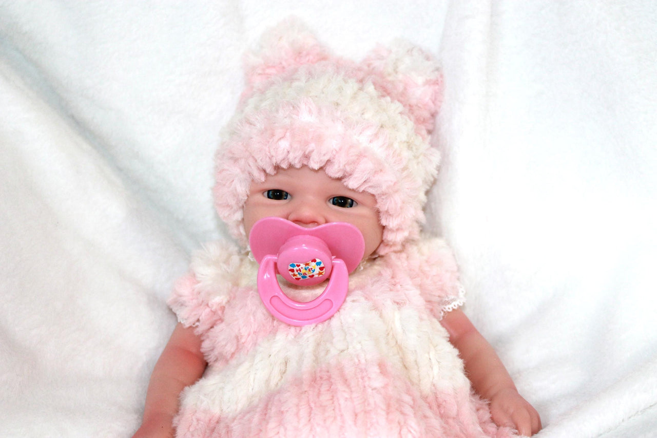 A baby doll dressed in a pink hat, featuring a pacifier, lying on a cozy blanket, highlighting its adorable design.