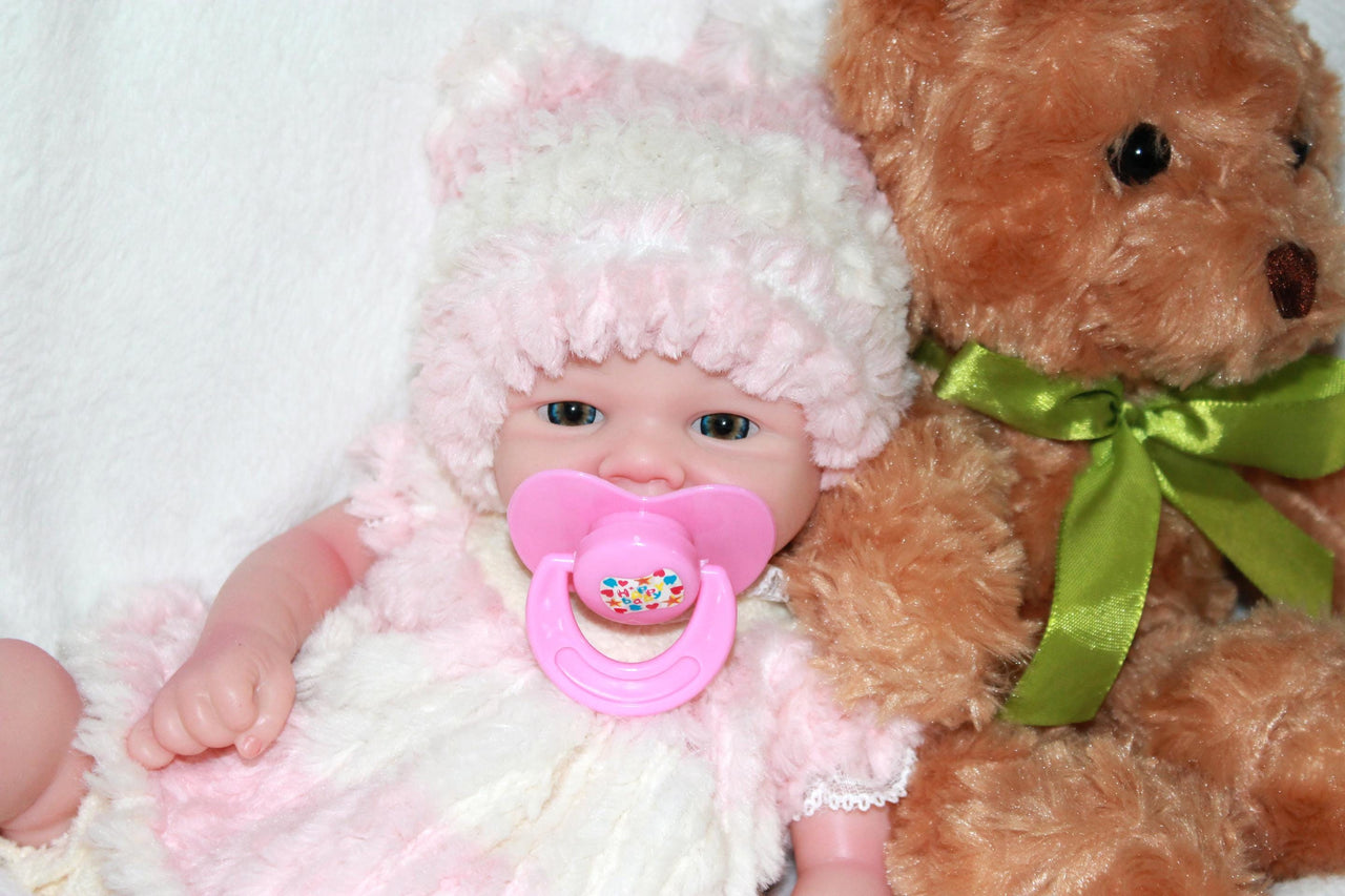 A baby doll with a pink hat and a pacifier, resting on a soft surface, showcasing its cute features.