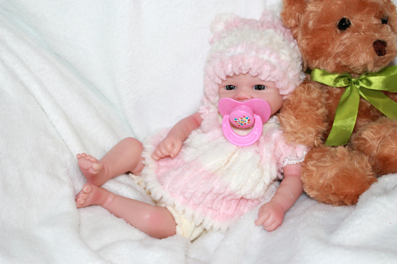 A baby doll dressed in a pink hat, featuring a pacifier, lying on a cozy blanket, highlighting its adorable design.