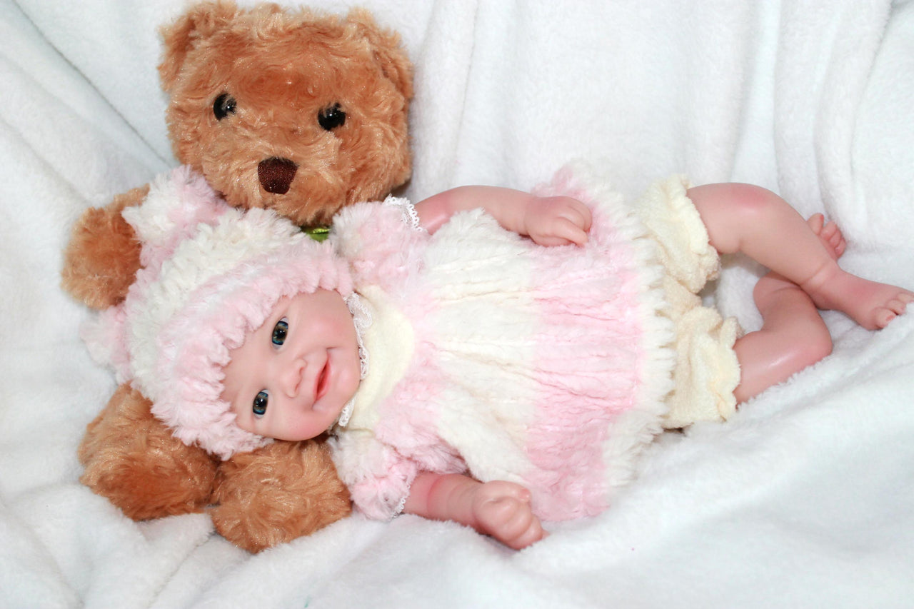 A baby doll dressed in a pink hat, featuring a pacifier, lying on a cozy blanket, highlighting its adorable design.