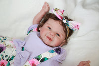 Thumbnail for A baby doll dressed in a purple dress and adorned with a flower headband.