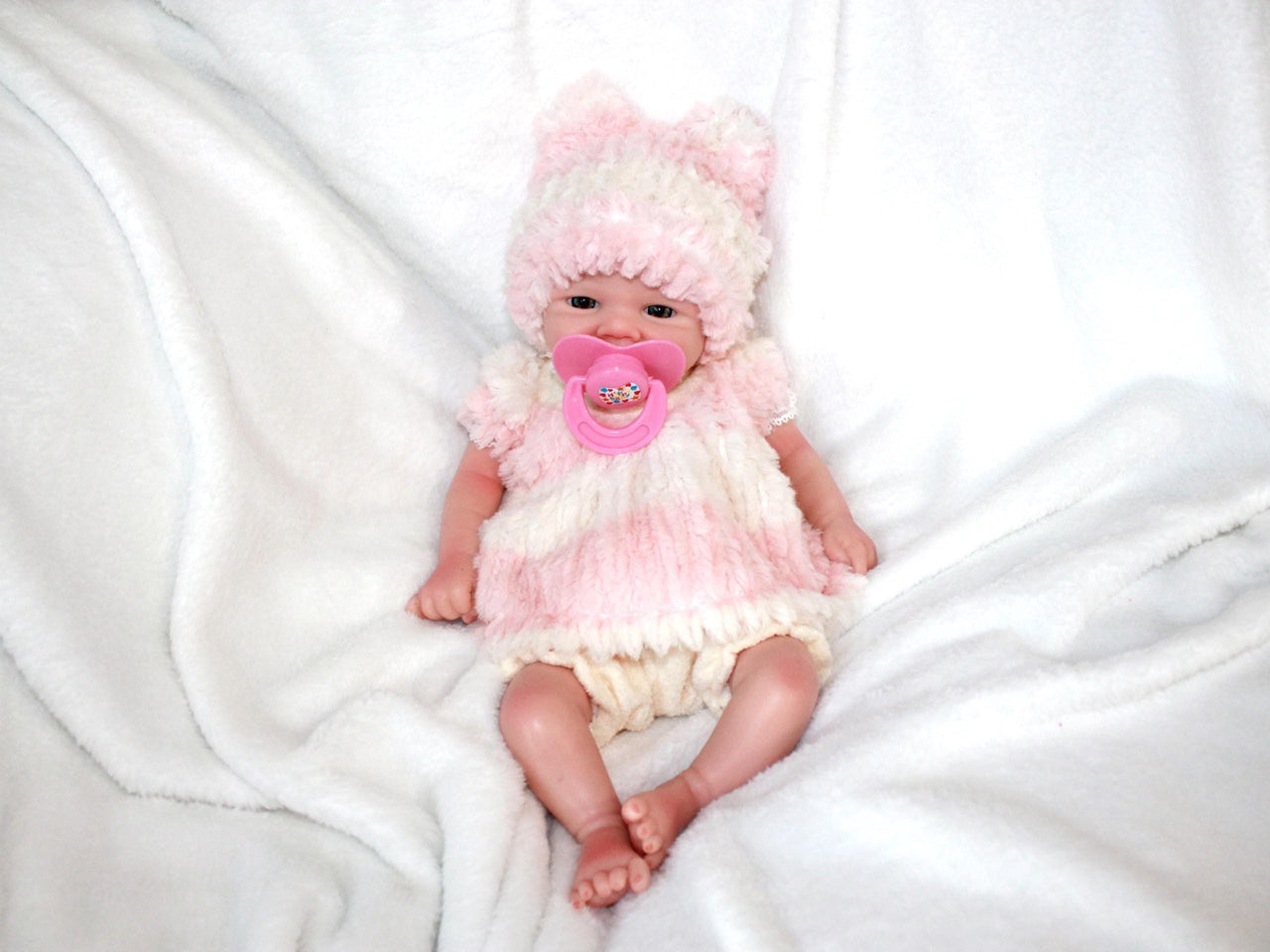 A baby doll dressed in a pink hat, featuring a pacifier, lying on a cozy blanket, highlighting its adorable design.