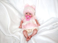 Thumbnail for A baby doll dressed in a pink hat, featuring a pacifier, lying on a cozy blanket, highlighting its adorable design.