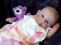 Thumbnail for A baby doll resting on a purple sheet, showcasing its soft features and delicate clothing.