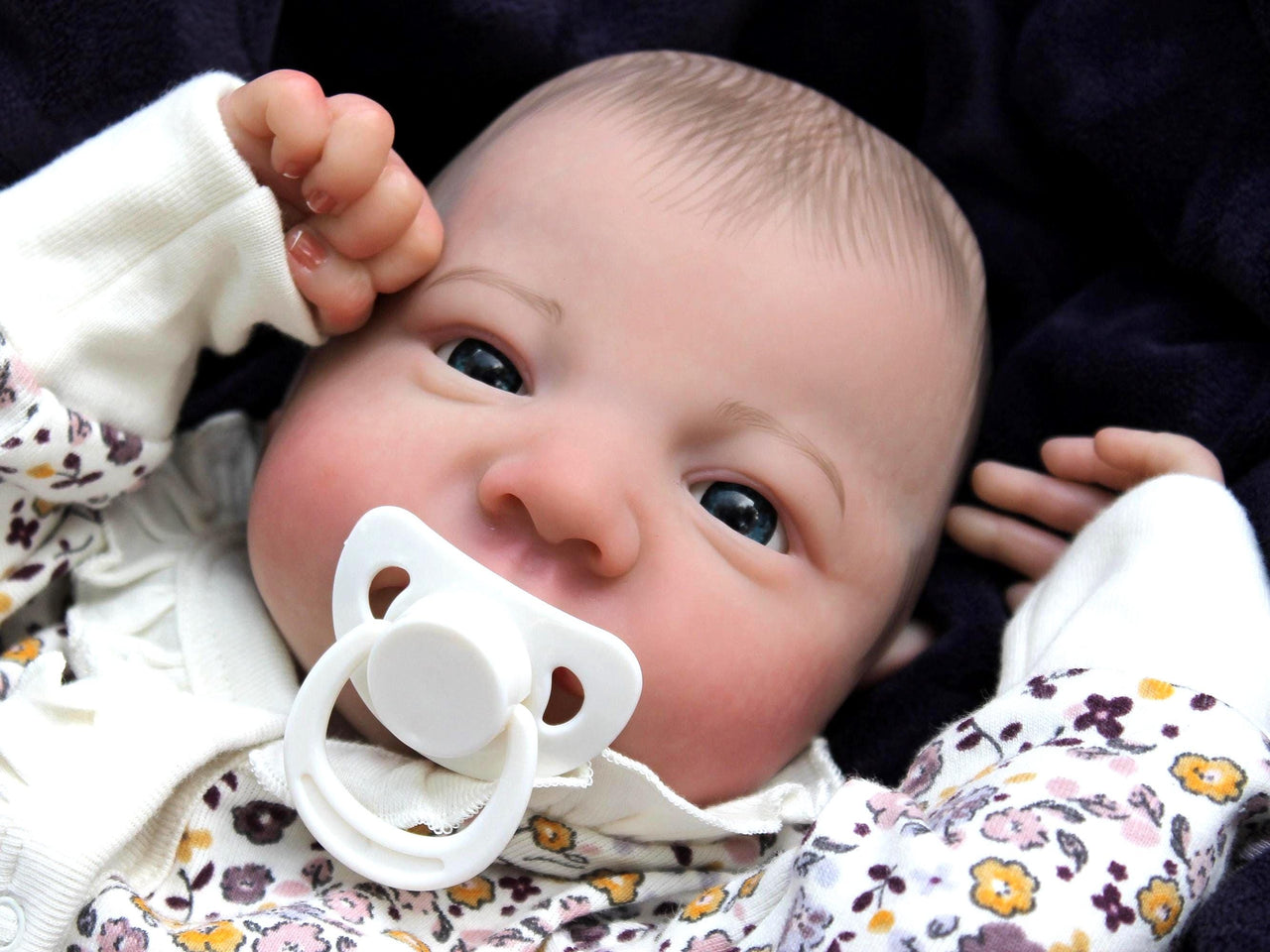A baby doll with a pacifier in its mouth, featuring soft facial features and a floral outfit.