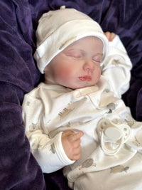 Thumbnail for A baby doll rests on a soft blanket, showcasing its delicate features and clothing.