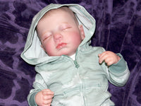 Thumbnail for A baby resting on a bed, adorned with a charming hat, surrounded by cozy blankets.