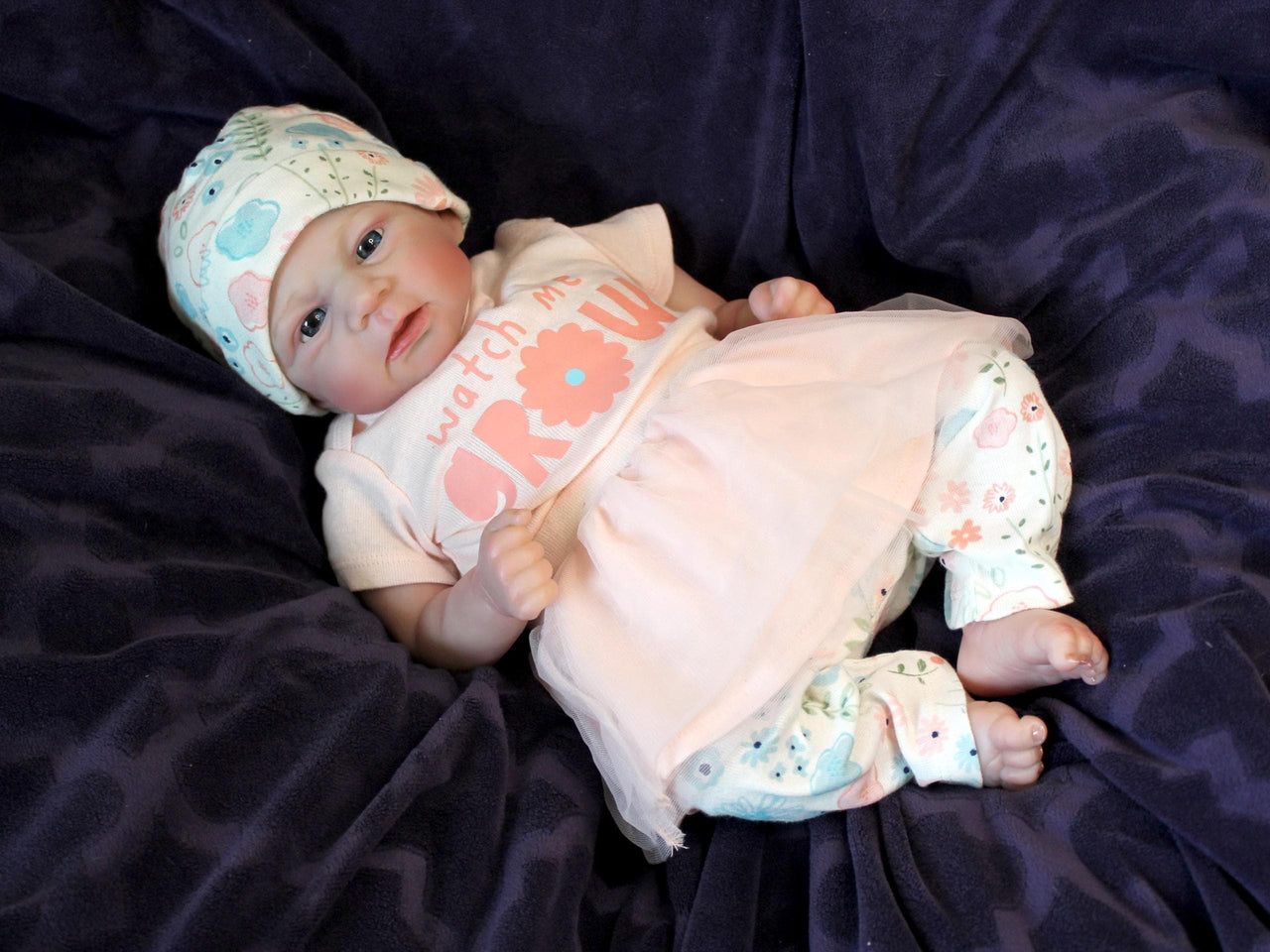 A baby doll dressed in a pink shirt and wearing a hat, sitting against a soft background.