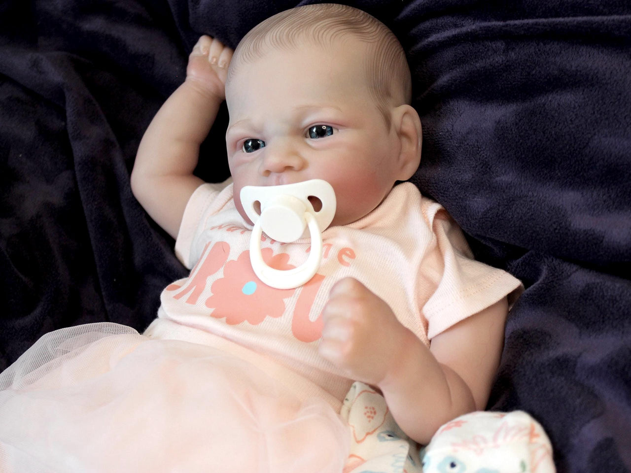 A baby doll with a pacifier lies on a cozy blanket, highlighting its charming design and soothing presence.