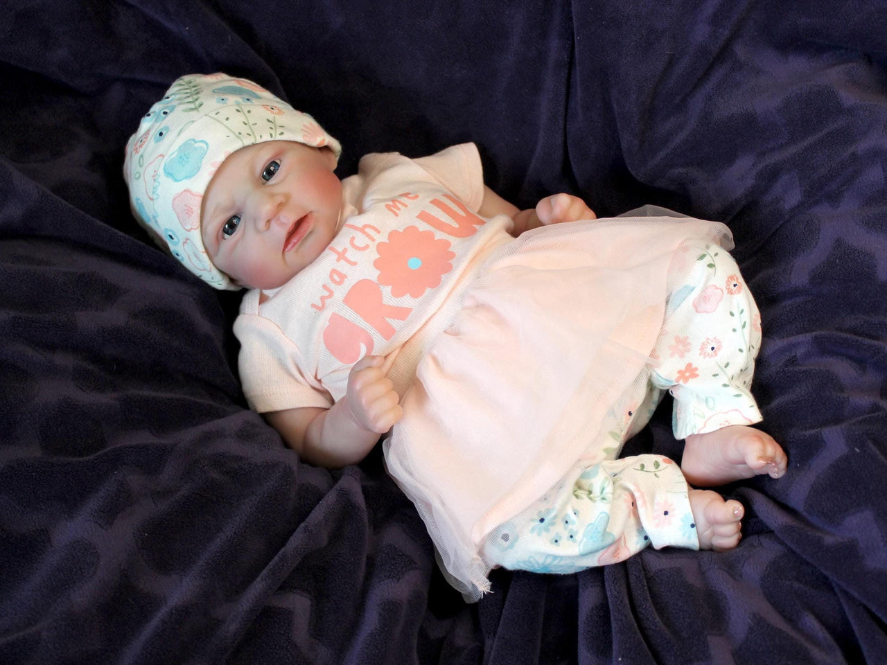A baby doll in a pink dress and hat, posed cutely with a gentle smile, highlighting its charming design.