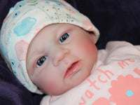 Thumbnail for A baby doll in a pink dress and hat, posed cutely with a gentle smile, highlighting its charming design.