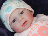 Thumbnail for A baby doll in a pink dress and hat, posed cutely with a gentle smile, highlighting its charming design.
