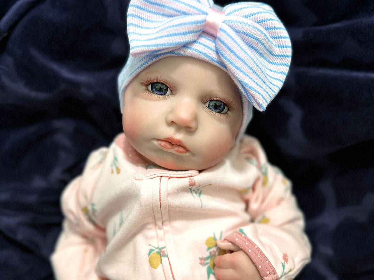 Lifelike reborn baby doll with blue eyes wearing a pink floral outfit and striped bow hat, realistic weighted newborn therapy doll