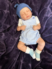 Thumbnail for Sleeping reborn baby doll wearing blue knit hat, blue checkered romper, and white socks on purple blanket – realistic handmade newborn doll