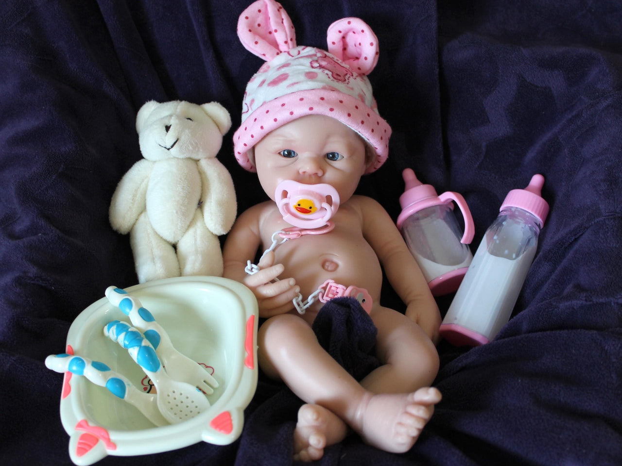Lifelike reborn baby doll with blue eyes wearing pink polka dot outfit and bunny ear hat, realistic handmade newborn doll on blanket