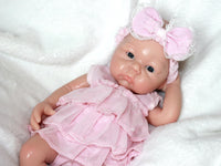 Thumbnail for A baby doll in a pink dress and headband, showcasing a cheerful expression and delicate features.