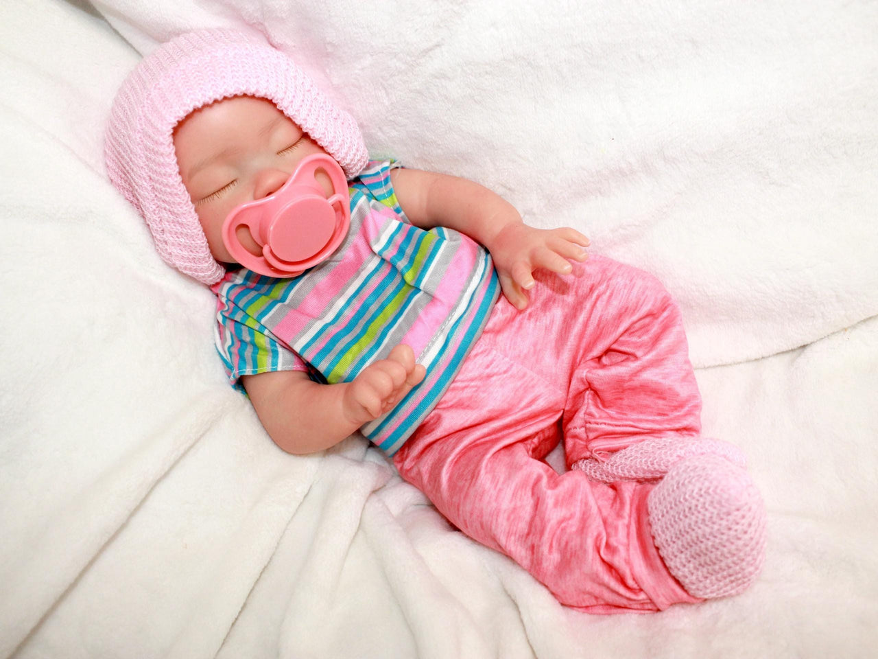 Sleeping reborn silicone baby doll wearing pink knit hat, striped shirt, and pink pants, lifelike 13-inch newborn doll on white blanket