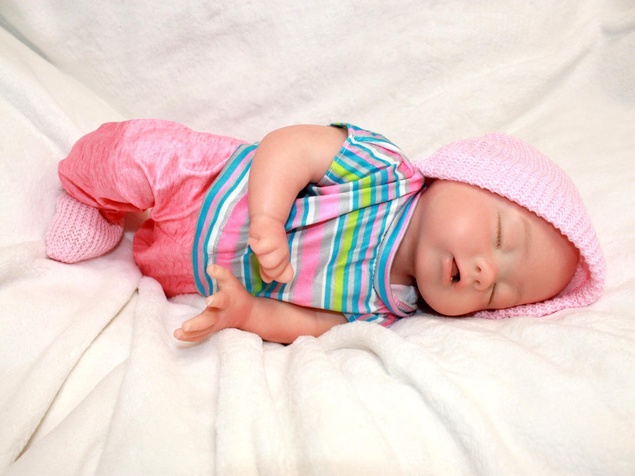 Sleeping reborn silicone baby doll wearing pink knit hat, striped shirt, and pink pants, lifelike 13-inch newborn doll on white blanket